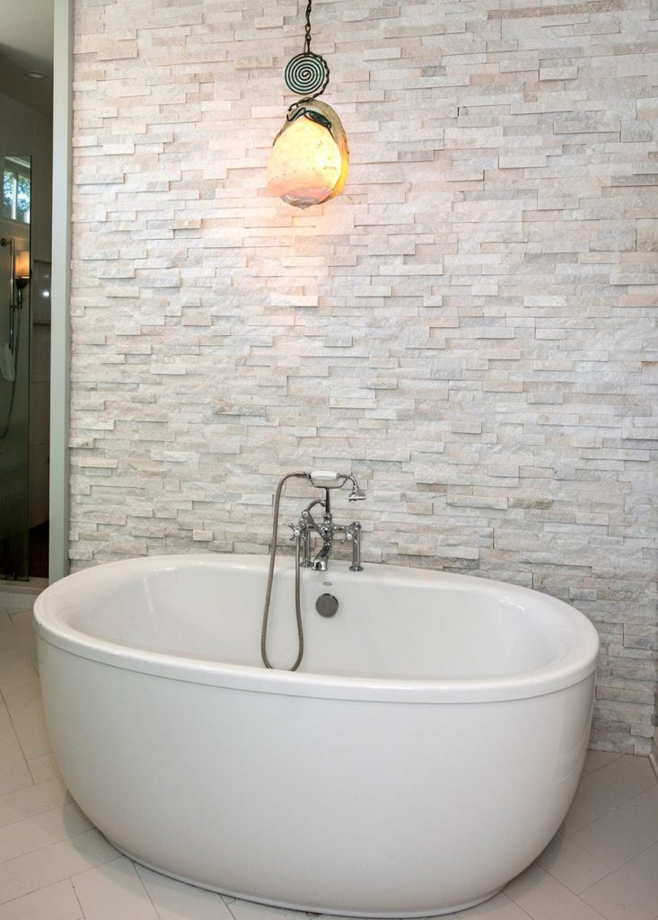 This large freestanding bathtub against a stone wall creates a mediterranean bathroom design in this Charleston interior design project by Provenance Lee Interiors